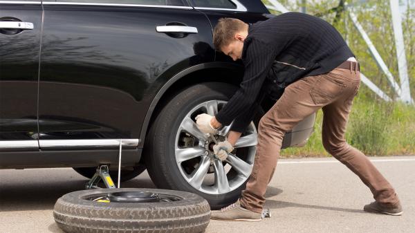 A young man turning a lug wrench on the lug bolts on the back tire of his black car.