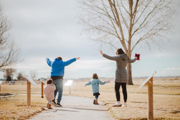 Two adults and two young girls with their arms wide open walking along a StoryWalk path.