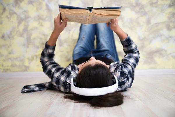 Girl laying on her back reading a book while wearing white headphones.