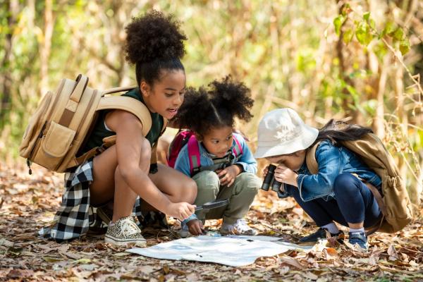 Three young girls look at a map using binoculars and a magnifying glass. They are surrounded by woods and dried leaves.