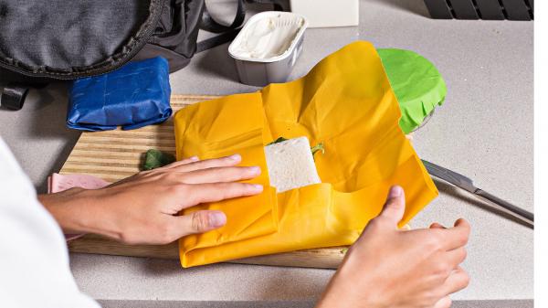 Two hands wrapping a sandwich in a bright yellow reusable beeswax wrap.