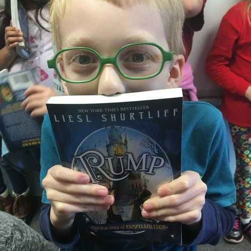 Child with green glasses holding juvenile book Rump by Liesl Shurtliff.
