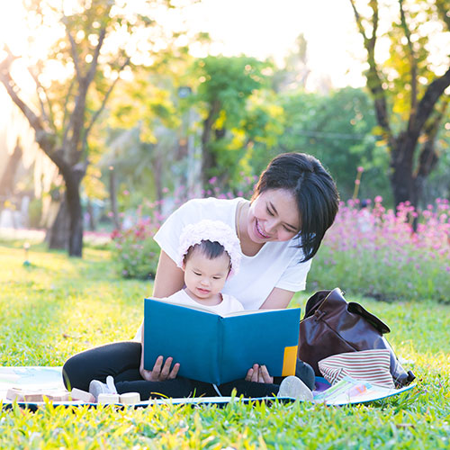Adult and older infant reading a book in the grass with flowers and trees behind them