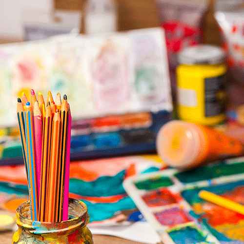 Table filled with colored pencils in a cup, tray of paint colors, paint in a tube and a jar, and blurry artwork in the background