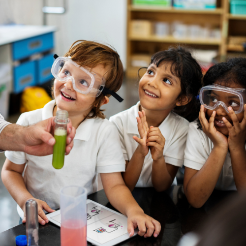 Three children smiling and looking at an adult holding a test tube, two children are wearing safety goggles.