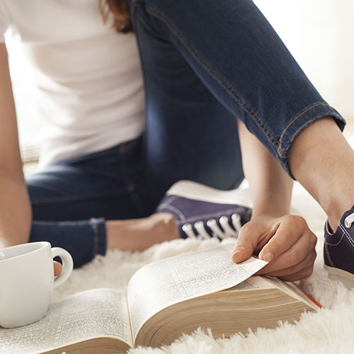 Girl sitting on the floor reading a book and holding a white coffee mug. 
