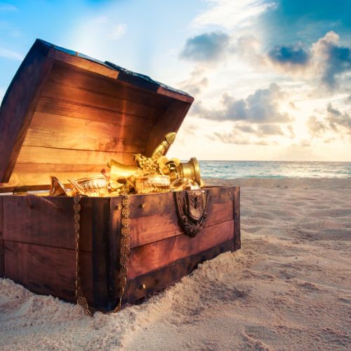 Open wooden chest filled with gold treasures sitting on the beach near the ocean.