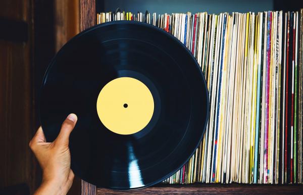 A hand holding a vinyl record with a yellow center in front of a shelf filled with records.